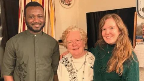 A young man, an older woman and a young woman stand together smiling with their arms around each other in a living room 