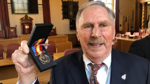 JOHN DEVINE/BBC Alan Dowson, who has short grey hair and a mustache, smiling into camera holding up Nuclear Test Medal. He is wearing a navy suit, white shirt and red-patterned tie. 