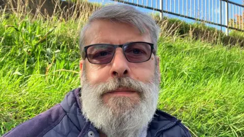 BBC/RICHARD EDWARDS Russell Dodgson looks directly into the camera. He has short grey hair and a grey beard, is wearing dark glasses and a blue jacket and is standing in front of a grassy bank.