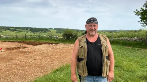 A man stood at an archaeological site wearing a khaki sleeveless jacket and jeabs. He has a bandana and goatee beard.