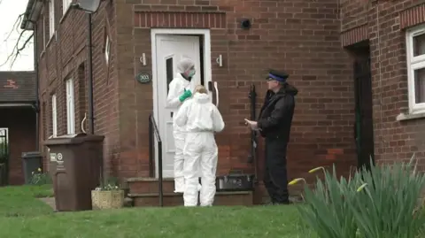 BBC Policeman in black uniform and cap speaks to two forensic officers in white kit as they stand outside the entrance to the brick house. A garden lawn and plants are at the front with wheelie bin at the side of the house.