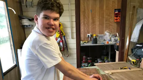 Family photo of Fraser in his dad's workshop holding a tool used to sand down his sailing boat.