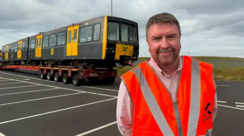 Paul Welford is standing in front of an original black and yellow Metro train. Welford has short grey hair and beard. He is wearing a pale pink shirt and a brigh orange high visibility vest.