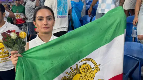 Supplied A young woman holds a bunch of roses and an Iranian green, white and red flag with the sun and the lion