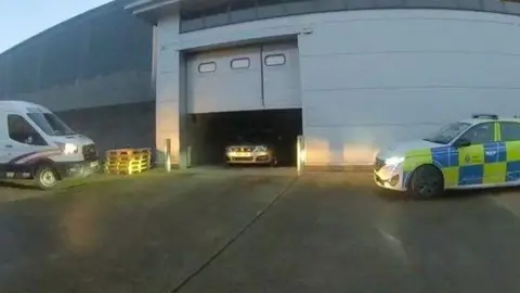 A business unit in High Brooms, Tunbridge Wells, where 3,500 cannabis plants were found. A white van is parked to the left of an open garage door, and saloon car is parked in the garage entrance and a police patrol car is parked to the right.