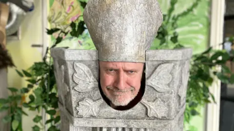 Bearded man with a bishops hat on poses with a hard stare inside a cut out would be stone pillar. The pillar is painted grey with leaves and is made of cardboard. 