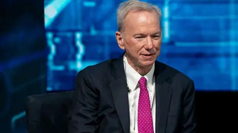 Eric Schmidt sits with his hands crossed and looks ahead. He is wearing a black suit, a white shirt, and a hot pink polka-dot tie. In the background is a blue screen that features different shades of blue.