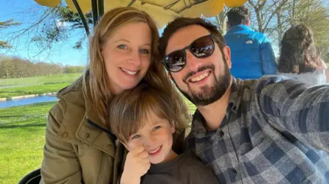 A woman and a child are sitting underneath a canopy as a man takes a selfie with them. The man is wearing a blue and grey chequered shirt and sunglasses. The woman is wearing a green coat. The child is wearing a grey t-shirt. All three are smiling and there is a body of water and a field behind them.