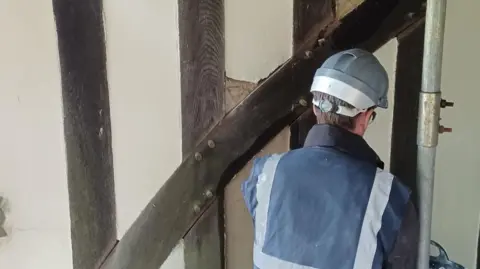 National Trust A man in a navy work jacket with fluorescent white strips on the black, wearing a hard hat. He is crouched with his back to the camera and appears to be working on a cream and timber-framed wall.
