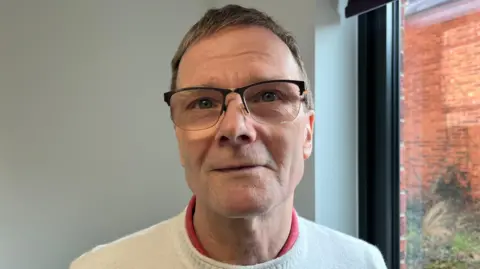 Patrick looking into the camera and smiling slightly; he's wearing a cream knitted jumper with a light red shirt underneath. He's stood by a window looking out onto a red brick courtyard with some greenery by the far wall. He's pictured inside against a white wall.