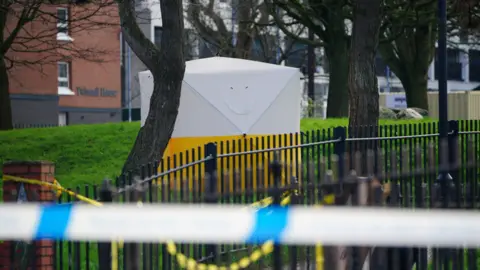 A forensic tent erected by metal railings with red brick flats visible in the background.  