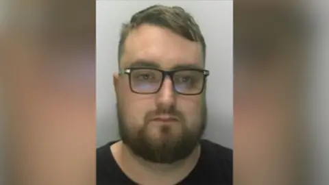 South West ROCU A mugshot of a man in his early 30s with short brown hair, which is pushed to the side, black framed glasses, and brown facial hair. He is wearing a black round neck T-shirt.