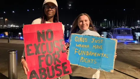 Mia Brown and Lauren Searle stand on a waterfront, with boats behind. It is night. They both hold banners, one reads No Excuse for Abuse, the other says Girls just wanna have fundamental rights