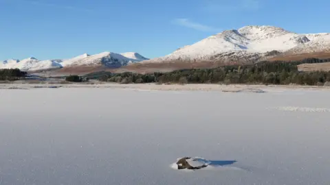 Gary Stanton Frozen loch with a boulder breaking through the sheet of ice with snow covered hills in the distance