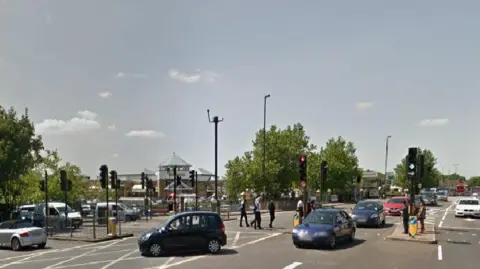 A road busy junction with a shopping centre in the background. People are crossing at the traffic lights and a queue of cars are waiting to turn right. 