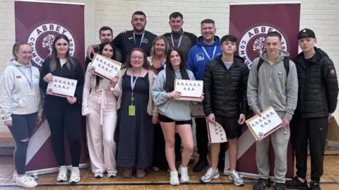 Monkstown Boxing Club A group of young people and adults standing in two lines, one in front of the other. They are all smiling. Some of the young people are holding up signs which  show their GCSE results in large letters. Behind them are vertical maroon banners which say Abbey Community College.  