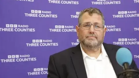 Peter Geary wearing a dark jacket and light-coloured shirt is seated in a radio studio in front of a microphone with a blue windscreen. The background is a purple branded backdrop repeating the text “BBC Radio Three Counties”. A second person is partially visible at the right edge of the image. The photograph is brightly lit and appears to have been taken during a radio interview.