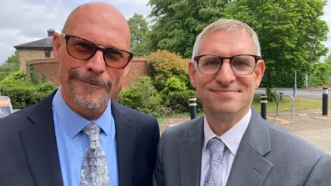 BBC Two men are looking at the camera, wearing suits. One is wearing a grey suit with a pink shirt and purple tie, while the other man is wearing a dark blue suit, blue shirt and silver patterned tie. Behind them is a council building.