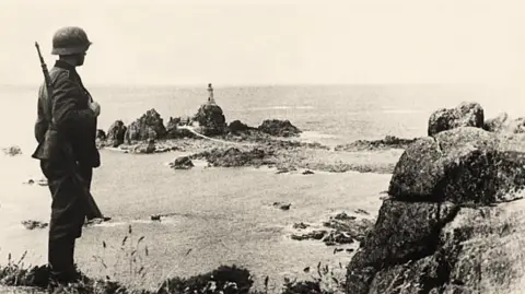 A German solider holding a rifle and wearing a tin helmet is standing on a cliff and looking out to sea