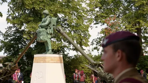 LAURA FOSTER/BBC The life-size statue is green and standing on a plinth. There are cannons on either side with troops seen standing in the background.