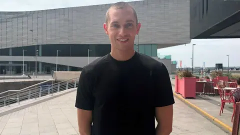 Sonny Walker, who is wearing a black T-shirt, is smiling at the camera as he standing on terrace outside a restaurant. 