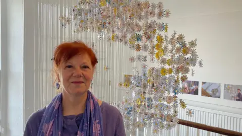 Gillian Taylor, who has red hair and is wearing purple, stands in front of her art installation. The daisies made from the letters hang on fishing wire from the ceiling of the stairwell.