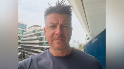 Luke Deal in a selfie-style image while he stands on a hotel balcony. He is slightly smiling at the camera. He has short dark hair with some grey. He wears a grey T-shirt.