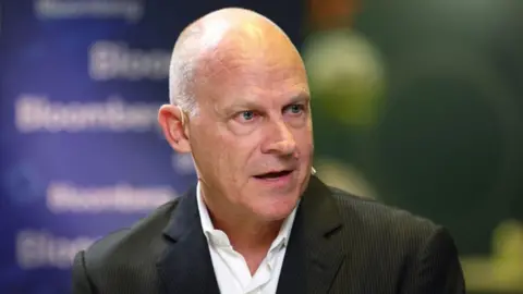 Campbell Wilson, wearing a black suit and a white shirt, speaks at a conference with a blue board behind him