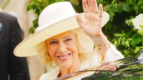 PA Media The Queen is pictured wearing a hat, holding her hand up at people to wave and is smiling
