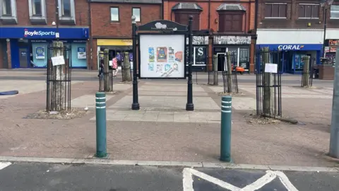 An empty town centre high street with several tree stumps encased in small black metal railings. There are shop fronts in the background.