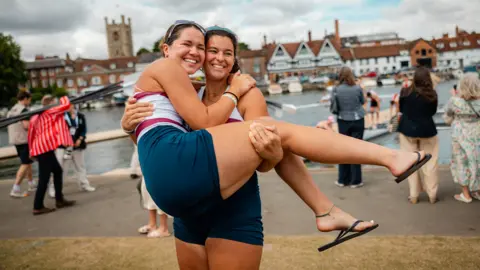 Henley Royal Regatta / Row360 A woman holding another woman as they pose for a picture on the first day of Henley Royal Regatta 2026. They are smiling. They are other people behind them. It is overcast.