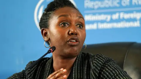PA Rwanda government spokesperson Yolande Makole sits on a black chair before a microphone. She is wearing a white dotted black blazer.