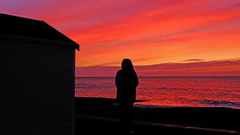 SQUIZ/WEATHERWATCHERS A silhouetted figure stands in front of the sea and next to what appears to be a beach hut, as an orange and red sunrise colours the sky