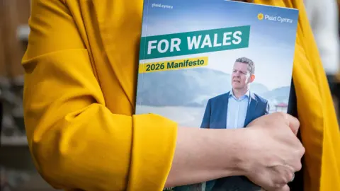 A woman in a yellow jacket holding a copy of the Plaid Cymru manifesto, which reads "for Wales" in capital letters and "2026 manifesto". The cover of the manifesto shows Rhun ap Iorwerth looking off into the distance.