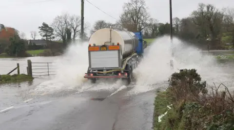El vehículo cisterna Pacemaker, con parte trasera plateada y cabina azul, salpica agua en una carretera cerca de una valla, con árboles al fondo.