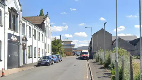 Vehicles are parked on either side of the street and a row of white buildings can be seen on the left.
