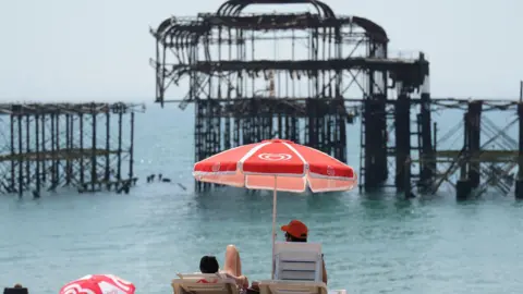 Eddie Mitchell Two people lying on a beach chair and sitting under a red umbrella. They are looking towards the metal structure of a former pier. 