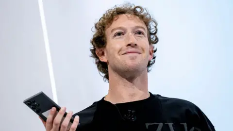 Getty Images A smiling Mark Zuckerberg stands on stage holding up a smartphone