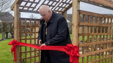Stoke-on-Trent City Council Lord Mayor of Stoke-on-Trent, Steve Watkins is stood under a wooden trellis with a red ribbon attached. He is holding a pair of scissors, and is cutting the ribbon.