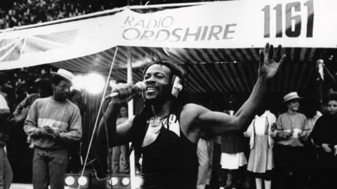 The Man Ezeke singing into a microphone in front of a tent outside. Behind him people are joining in with the music. He has his arm in the air and his eyes closed.