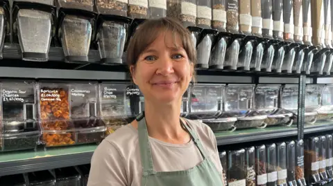 Ellen Knight/BBC Marie MacNae pictured looking into the camera and smiling. She is wearing a beige t-shirt, with a light green apron over the top. She is stood inside her refill shop, with tubs of dried fruit and nuts behind her. 