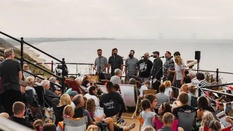 Saltburn Smugglers Eight performers stand in a line on the promenade and sing into microphones. There is a crowd of people on the stairs and in deckchairs watching on. The outline of a wind farm on the water can be made out in the mist. The sandy coastline stretches out beyond them.