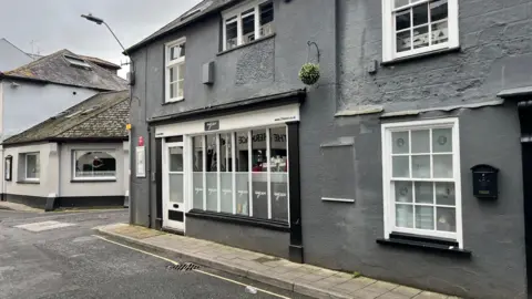 Grey painted two storey buidling with white wood-framed and sash windows. Part of the road and another building are also visible in the foreground and background and it is an overcast day.