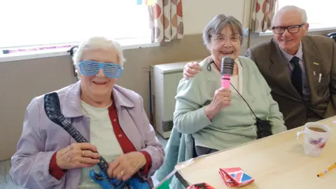 b:friend Three older adults sit at a table in a bright room. One person on the left wears novelty blue shutter-style glasses and holds a small blue toy guitar. The person in the centre holds a pink-handled microphone, while the person on the right, wearing glasses and a jacket, has an arm around the centre person. On the table are a mug, a pencil, and playing cards, suggesting a social or activity setting.