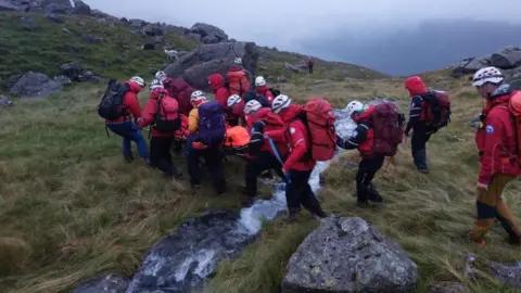 Wasdale Mountain Rescue Team Wasdale Mountain Rescue Team carrying the casualty on a stretcher. A group of several people are wearing red jackets and white helmets. The are crossing a small stream on a hillside.