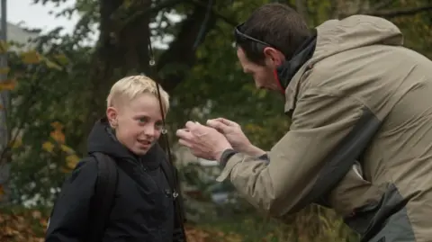 BBC Fisherman Kieron Banks prepares a rod and a line while a young boy, who has blonde hair, looks at the hook being prepared. They are standing by a river bank which has lots of green shrubbery. 