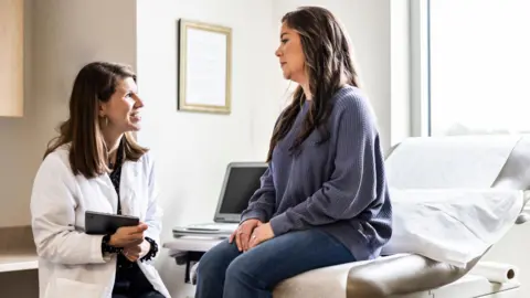 A Female doctor with mid length brown hair, wearing a white coat, holds an ipad  and discusses treatment with female patient who sits on a bed, and wears a blue jumper in an exam room