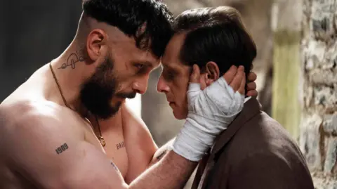 A man with a beard and bandages on his hands holds the face of a man wearing a suit.