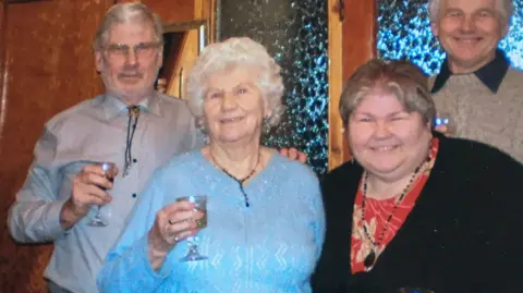 Willina, a resident of Whitestones care home, surrounded by her grown-up children, smiles in a blue jumper holding a drink