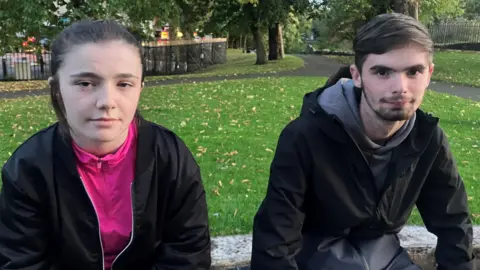 Kenzie and John are pictured sitting on a wall. Kenzie has brown hair tied back and is wearing a pink zip up jacket and a black zip up jacket over the top. John has dark brown hair and is wearing a grey hoodie and a black waterproof jacket on top. 